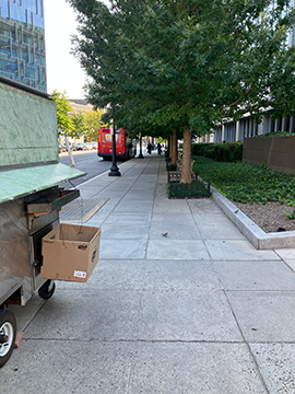 A picture of a sidewalk down 7th Street. The WMATA Headquarters building is on the left, and the street is on the right. There is landscaping and some construction cones to the right before the street.